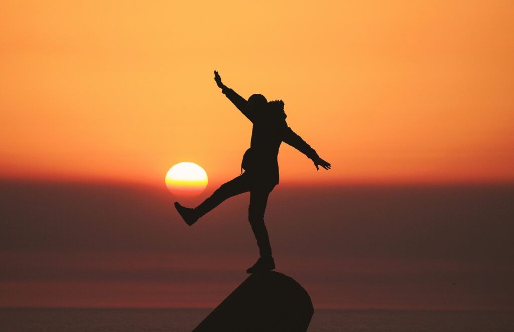 A silhouette of a person standing on a rock with their arms outstretched with the sun rising in the background.