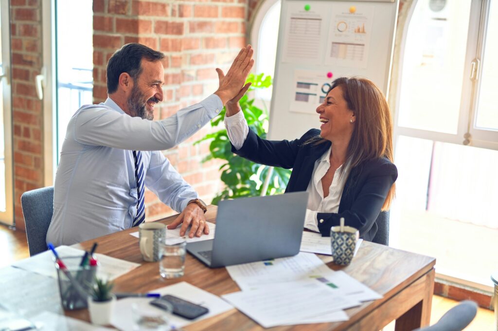 A man and woman in business attire sitting at a desk, smiling and high-fiving each other.